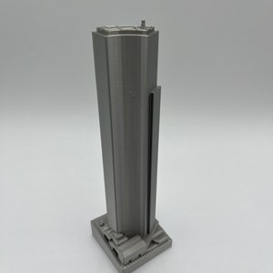 May include: A 3D printed model of a skyscraper in a silver gray color. The model has a rectangular base and a tall, slender tower with a flat roof.