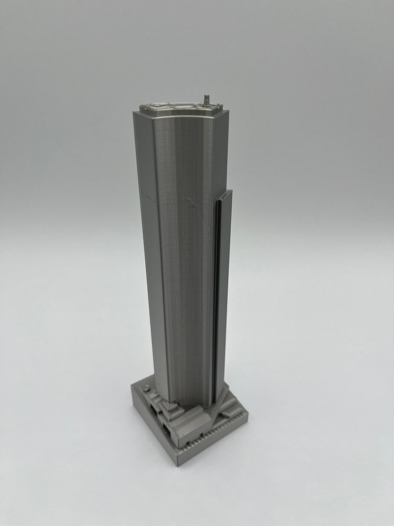 May include: A 3D printed model of a skyscraper in a silver gray color. The model has a rectangular base and a tall, slender tower with a flat roof.