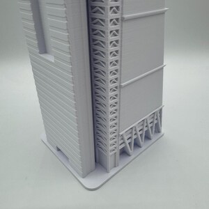 May include: A white architectural model of a skyscraper. The model features detailed facades, including horizontal lines and a decorative support structure. The building sits on a rectangular base, showcasing a modern design.