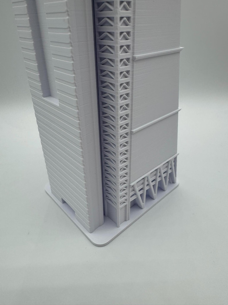 May include: A white architectural model of a skyscraper. The model features detailed facades, including horizontal lines and a decorative support structure. The building sits on a rectangular base, showcasing a modern design.