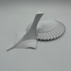 May include: A white 3D printed model of the Olympic Stadium in Montreal, Canada. The stadium is a large, circular structure with a distinctive tower.