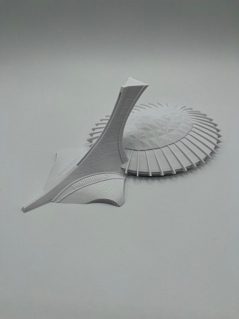 May include: A white 3D printed model of the Olympic Stadium in Montreal, Canada. The stadium is a large, circular structure with a distinctive tower.
