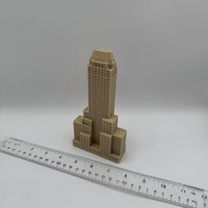 Może przedstawiać: Beżowy model miniaturowego budynku z detalami, przypominający wieżowiec. Przezroczysta linijka z oznaczeniami w centymetrach umieszczona przed budynkiem. Model budynku ma około 13 cm wysokości.