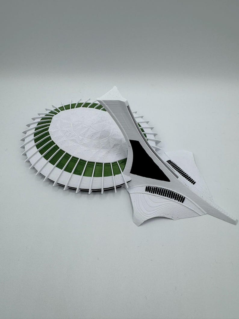 Puede incluir: Maqueta arquitect&oacute;nica blanca y verde. La base circular tiene una superficie blanca texturizada con detalles verdes. Una estructura en forma de ala se extiende desde la base, con detalles negros.