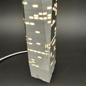 May include: A gray, 3D-printed skyscraper lamp with illuminated rectangular windows. The lamp has a textured surface and a white power cord. The windows emit a warm, yellow light, creating a cozy ambiance. The lamp is a modern home decor item.