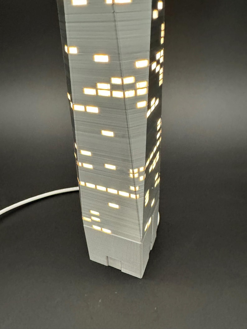 May include: A gray, 3D-printed skyscraper lamp with illuminated rectangular windows. The lamp has a textured surface and a white power cord. The windows emit a warm, yellow light, creating a cozy ambiance. The lamp is a modern home decor item.