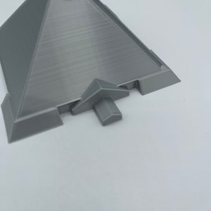 Puede incluir: Modelo de pir&aacute;mide gris impreso en 3D con una superficie texturizada. La pir&aacute;mide tiene una base plana y una peque&ntilde;a estructura que sobresale en un lado. El modelo est&aacute; sobre un fondo blanco.