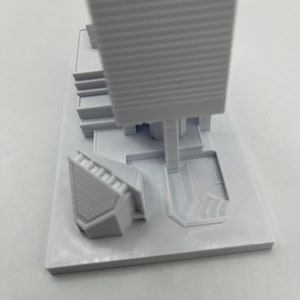 May include: White 3D printed model of a skyscraper with a smaller building and a plaza in front. The model is on a white base.