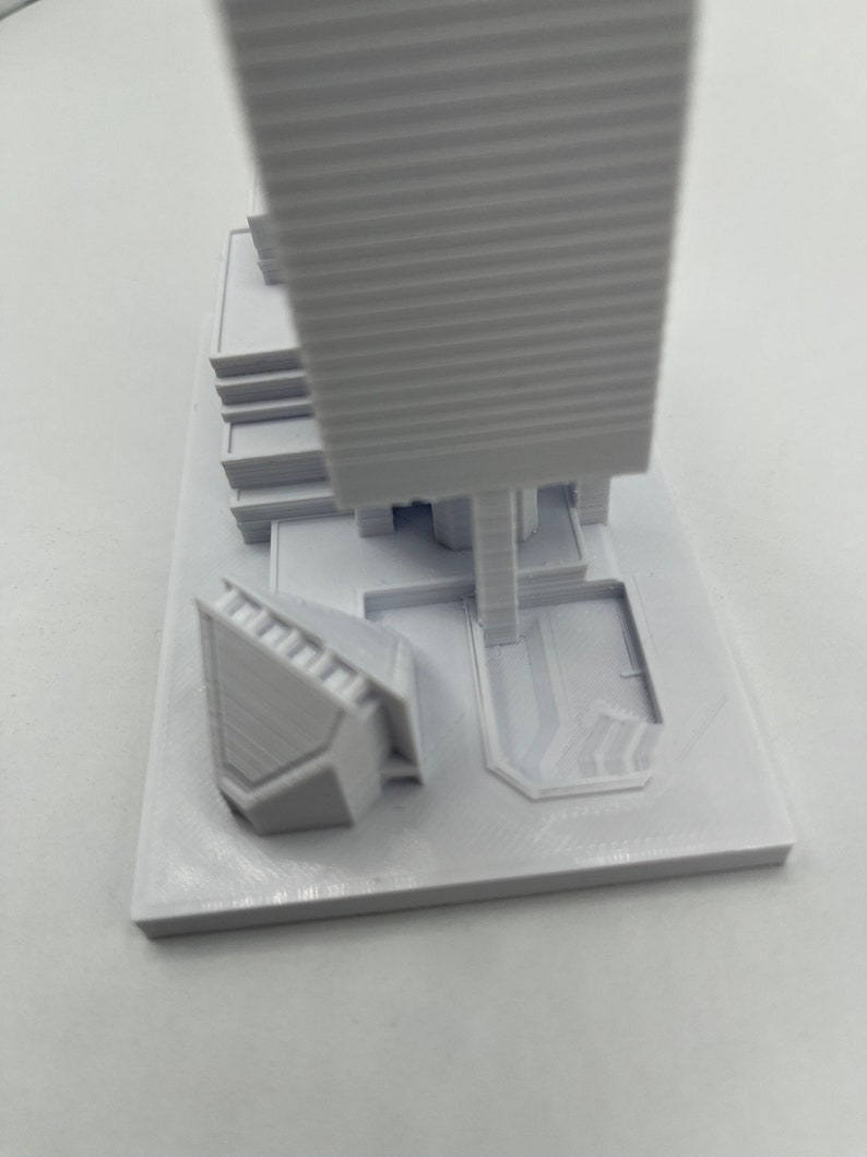 May include: White 3D printed model of a skyscraper with a smaller building and a plaza in front. The model is on a white base.