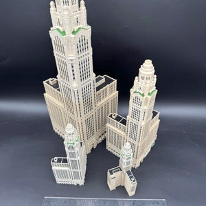 May include: Four off-white miniature skyscraper models with green accents on a black background. The models vary in size, with the tallest one prominently displayed. A ruler is placed in the foreground for scale.