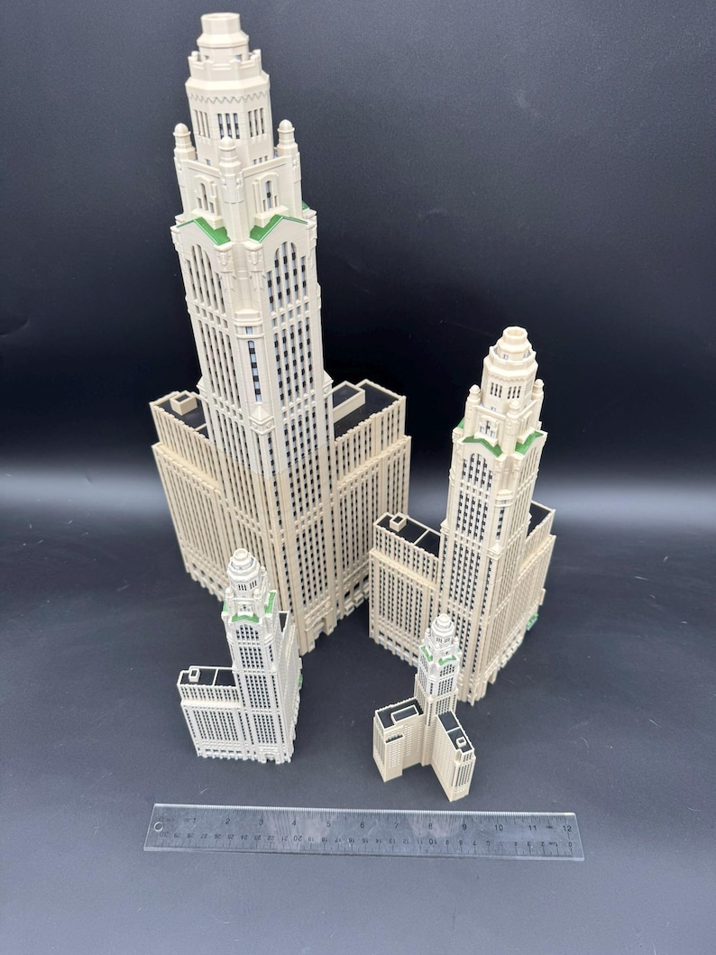 May include: Four off-white miniature skyscraper models with green accents on a black background. The models vary in size, with the tallest one prominently displayed. A ruler is placed in the foreground for scale.