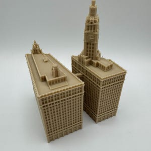 May include: Two beige miniature building models. One model is a tall skyscraper with a clock tower. The other model is a shorter building with a flat roof. Both models have detailed architectural features.