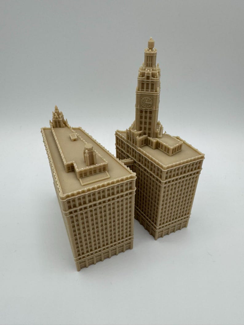 May include: Two beige miniature building models. One model is a tall skyscraper with a clock tower. The other model is a shorter building with a flat roof. Both models have detailed architectural features.