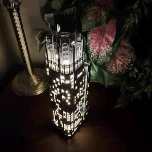 May include: A black and white detailed model of a skyscraper with lit windows. The model is on a dark surface with a gold lamp in the background.