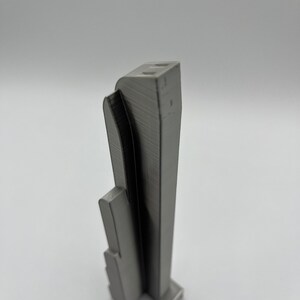 May include: A gray, 3D-printed architectural model of a skyscraper. The model has a rectangular base and a tall, slender tower with a curved facade. The building has a textured surface and appears to be made of plastic.