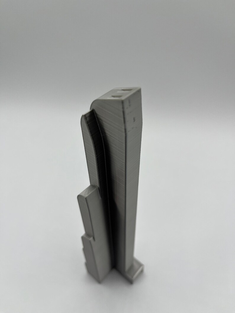 May include: A gray, 3D-printed architectural model of a skyscraper. The model has a rectangular base and a tall, slender tower with a curved facade. The building has a textured surface and appears to be made of plastic.