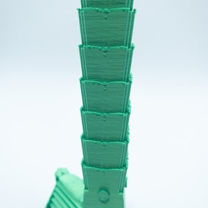 May include: A green 3D printed model of a tall building with multiple levels. The building has a rounded top and a small circular window.