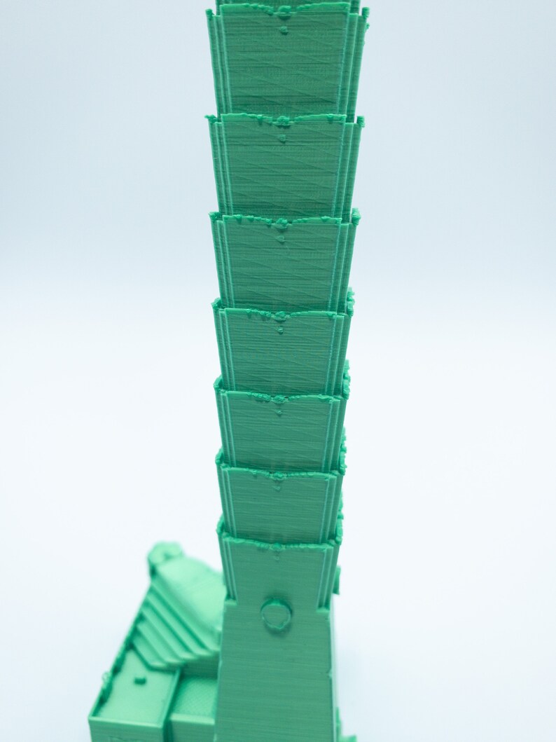 May include: A green 3D printed model of a tall building with multiple levels. The building has a rounded top and a small circular window.