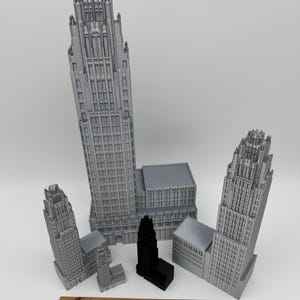 May include: A collection of gray and black miniature building models, including skyscrapers and other architectural structures. A wooden ruler is placed at the bottom of the image, providing a size reference. The models vary in size and design.