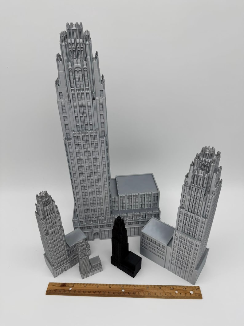 May include: A collection of gray and black miniature building models, including skyscrapers and other architectural structures. A wooden ruler is placed at the bottom of the image, providing a size reference. The models vary in size and design.
