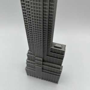 K&ouml;nnte beinhalten: Ein graues Miniaturmodell des Empire State Building. Das Modell hat eine detaillierte Fassade mit zahlreichen Fenstern und einem gestuften Design, das die Architektur des ikonischen Wolkenkratzers zeigt. Das Geb&auml;ude steht vor einem schlichten wei&szlig;en Hintergrund.