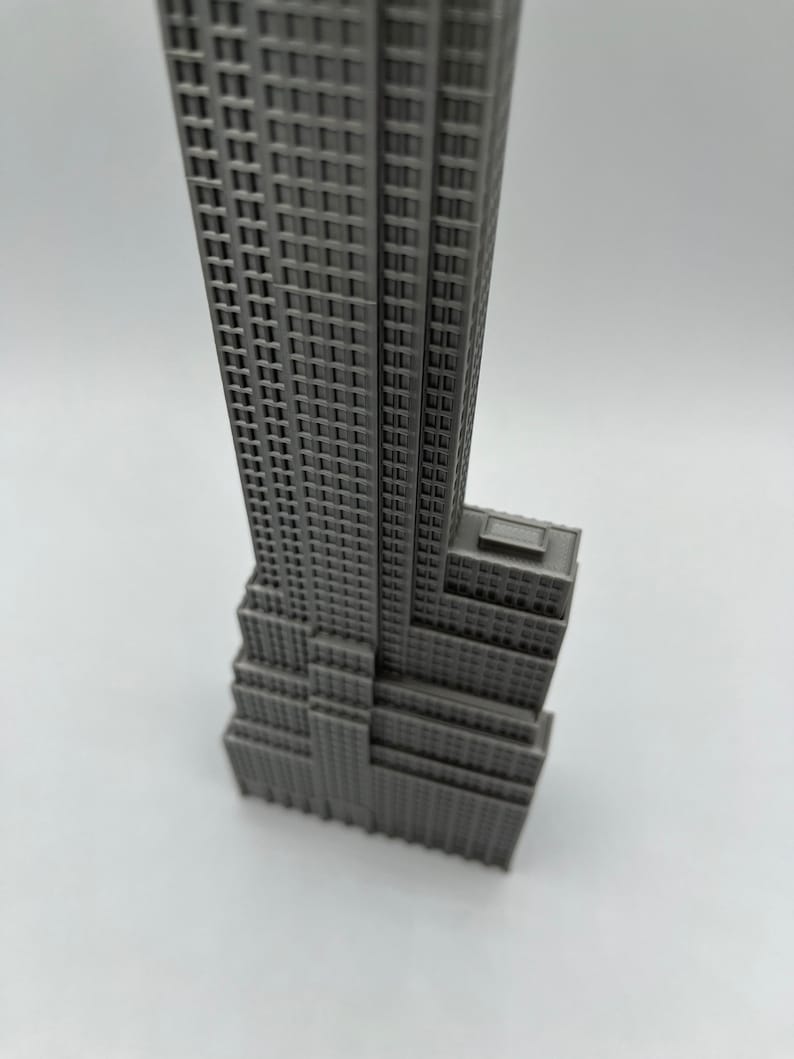 K&ouml;nnte beinhalten: Ein graues Miniaturmodell des Empire State Building. Das Modell hat eine detaillierte Fassade mit zahlreichen Fenstern und einem gestuften Design, das die Architektur des ikonischen Wolkenkratzers zeigt. Das Geb&auml;ude steht vor einem schlichten wei&szlig;en Hintergrund.
