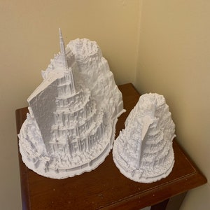 May include: Two white 3D printed models of a fictional city built on a mountain. The larger model has a tall tower at the top. The models are on a wooden surface.