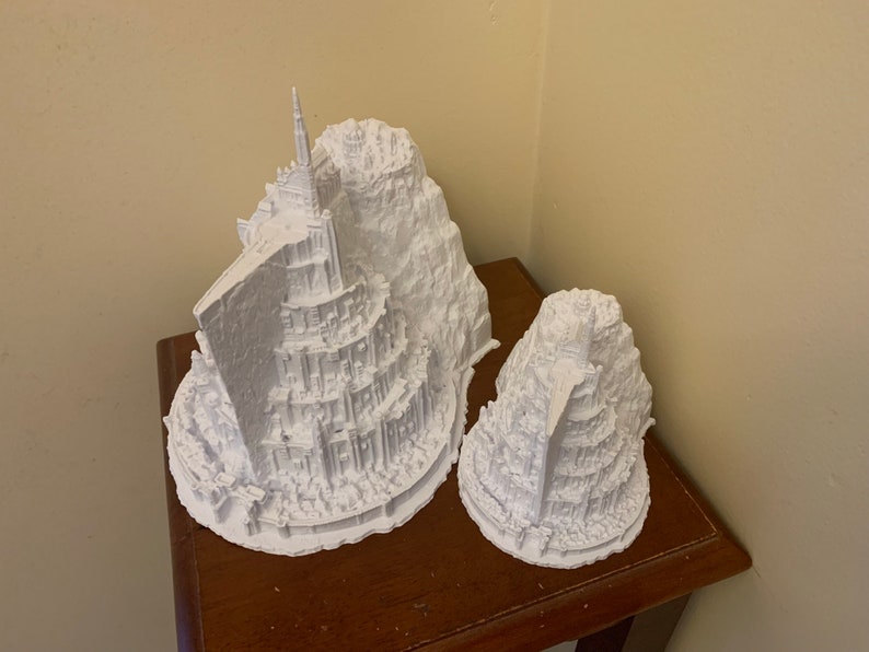 May include: Two white 3D printed models of a fictional city built on a mountain. The larger model has a tall tower at the top. The models are on a wooden surface.