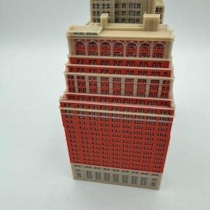 May include: A detailed model of a tall red brick building with many windows. The building has a flat roof and white trim around the windows. The model is made of plastic and is about 25 cm tall.