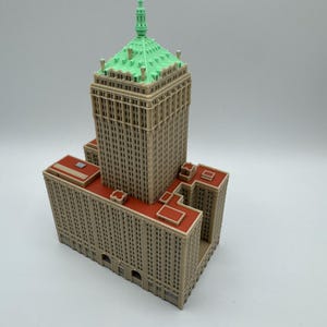 May include: A detailed miniature model of a skyscraper. The building features a tan facade with numerous windows, a red roof, and a green, pyramid-shaped top. The model is a detailed representation of a building.