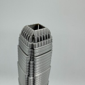 May include: A gray, 3D-printed model of a skyscraper. The building has a detailed, textured facade with horizontal lines and a square top. The model is a solid, architectural representation.