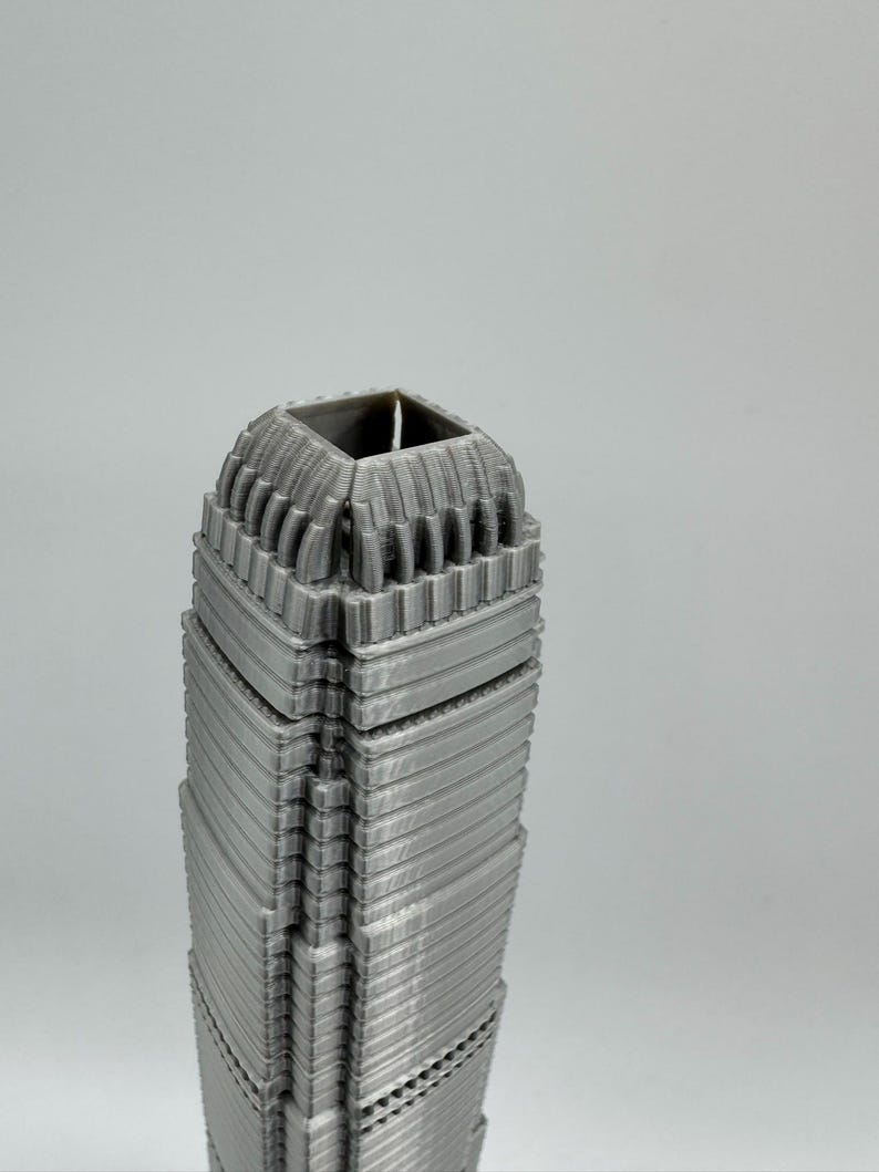 May include: A gray, 3D-printed model of a skyscraper. The building has a detailed, textured facade with horizontal lines and a square top. The model is a solid, architectural representation.