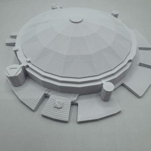 May include: A light gray architectural model of a domed structure with a circular base. The model features a detailed, layered design with a central dome and surrounding platforms. The overall design is symmetrical and geometric.