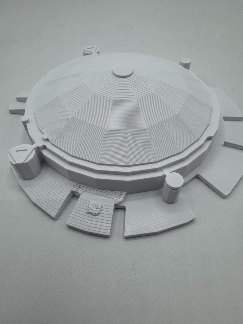 May include: A light gray architectural model of a domed structure with a circular base. The model features a detailed, layered design with a central dome and surrounding platforms. The overall design is symmetrical and geometric.