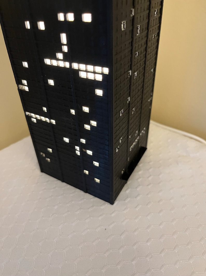 May include: A black model of a skyscraper with windows lit up, creating a pattern of light.
