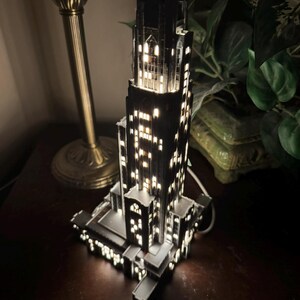 May include: A dark, multi-tiered skyscraper model lamp featuring intricate architectural details and numerous illuminated windows that emit a warm, bright glow from within the structure, creating a realistic night-time city building appearance.