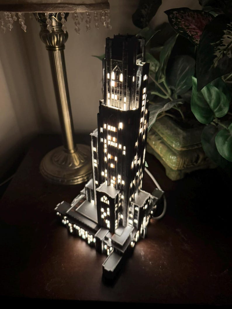 May include: A dark, multi-tiered skyscraper model lamp featuring intricate architectural details and numerous illuminated windows that emit a warm, bright glow from within the structure, creating a realistic night-time city building appearance.