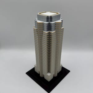 May include: A 3D-printed model of a skyscraper. The building is primarily light gray with white and dark gray accents. It has a rectangular base and a detailed facade, showcasing a modern architectural design.
