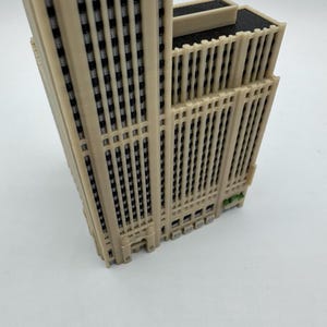 May include: A beige 3D-printed model of a skyscraper. The building has many windows and vertical lines. The top of the building is flat with a dark gray roof. The model is on a white surface.