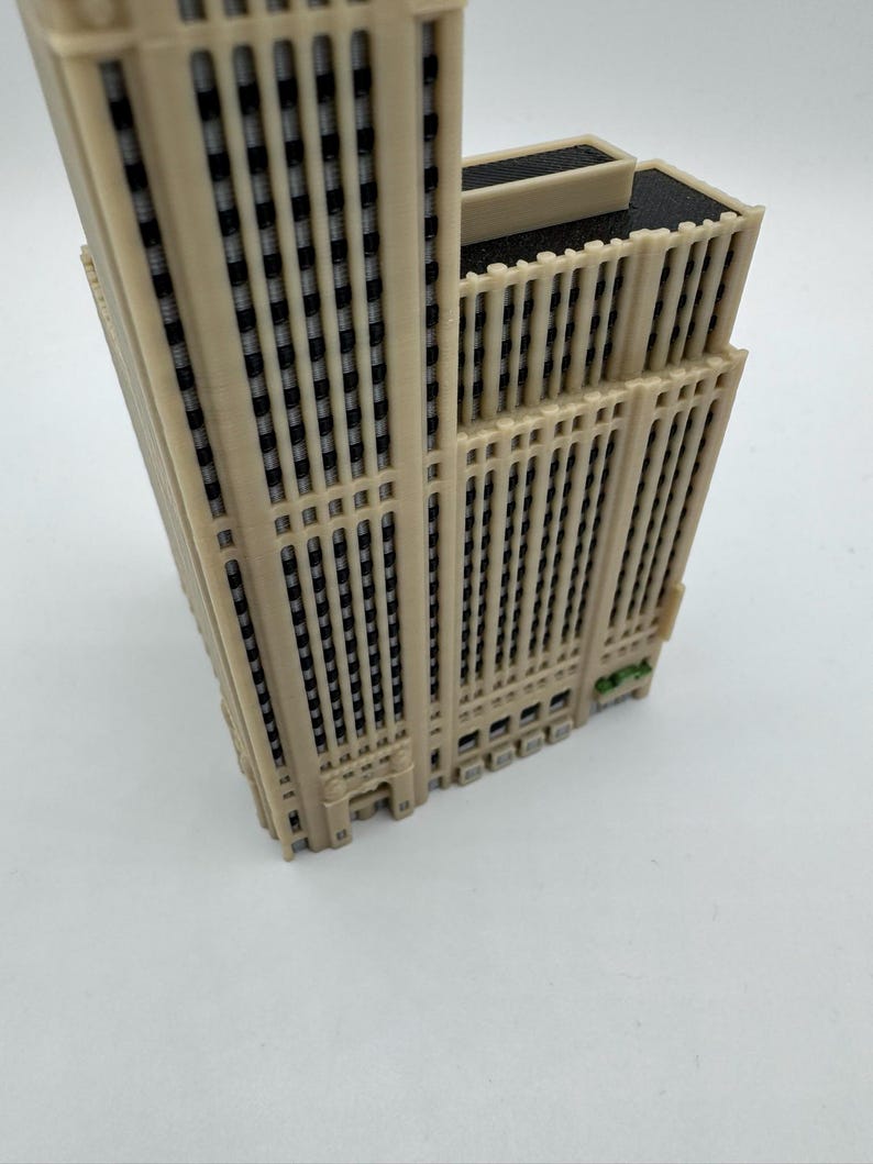 May include: A beige 3D-printed model of a skyscraper. The building has many windows and vertical lines. The top of the building is flat with a dark gray roof. The model is on a white surface.