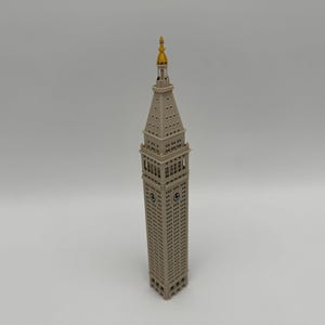 May include: A beige architectural model of the Metropolitan Life Insurance Company Tower, featuring a square clock tower base, a tapered spire, and a gold-colored pointed finial at the very top, captured from a high-angle perspective.