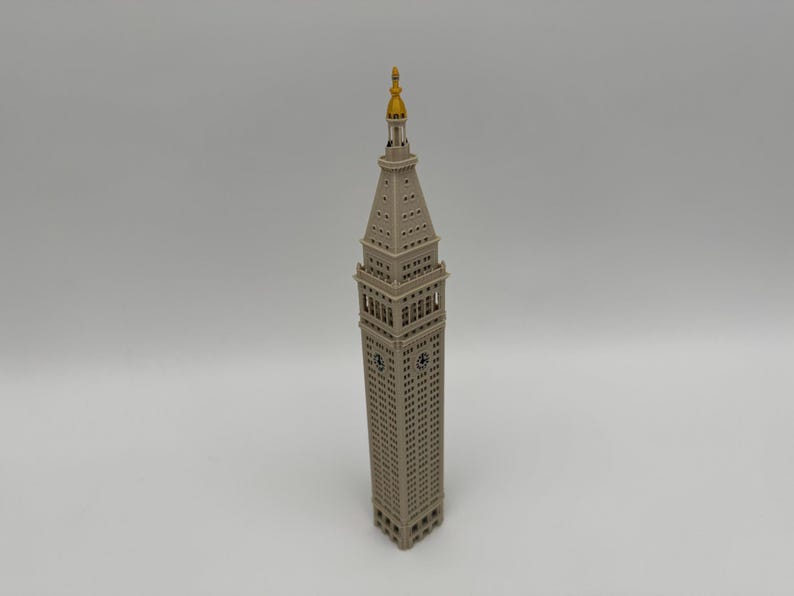 May include: A beige architectural model of the Metropolitan Life Insurance Company Tower, featuring a square clock tower base, a tapered spire, and a gold-colored pointed finial at the very top, captured from a high-angle perspective.