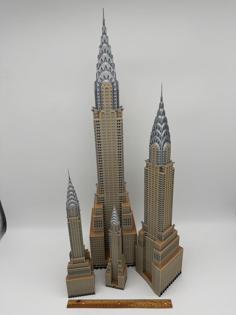 May include: A collection of five detailed miniature models of the Chrysler Building, featuring tan and silver color schemes. The models vary in size, showcasing the iconic Art Deco architecture. A wooden ruler is at the bottom.