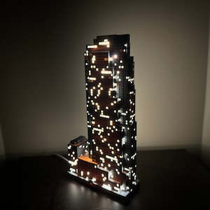 May include: A black and brown skyscraper model with white lights illuminating the windows. The model is lit from within and sits on a dark surface.
