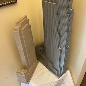 May include: Two 3D printed models of skyscrapers, one is gray and the other is beige. The models are on a white tablecloth with a ruler measuring 12 inches.