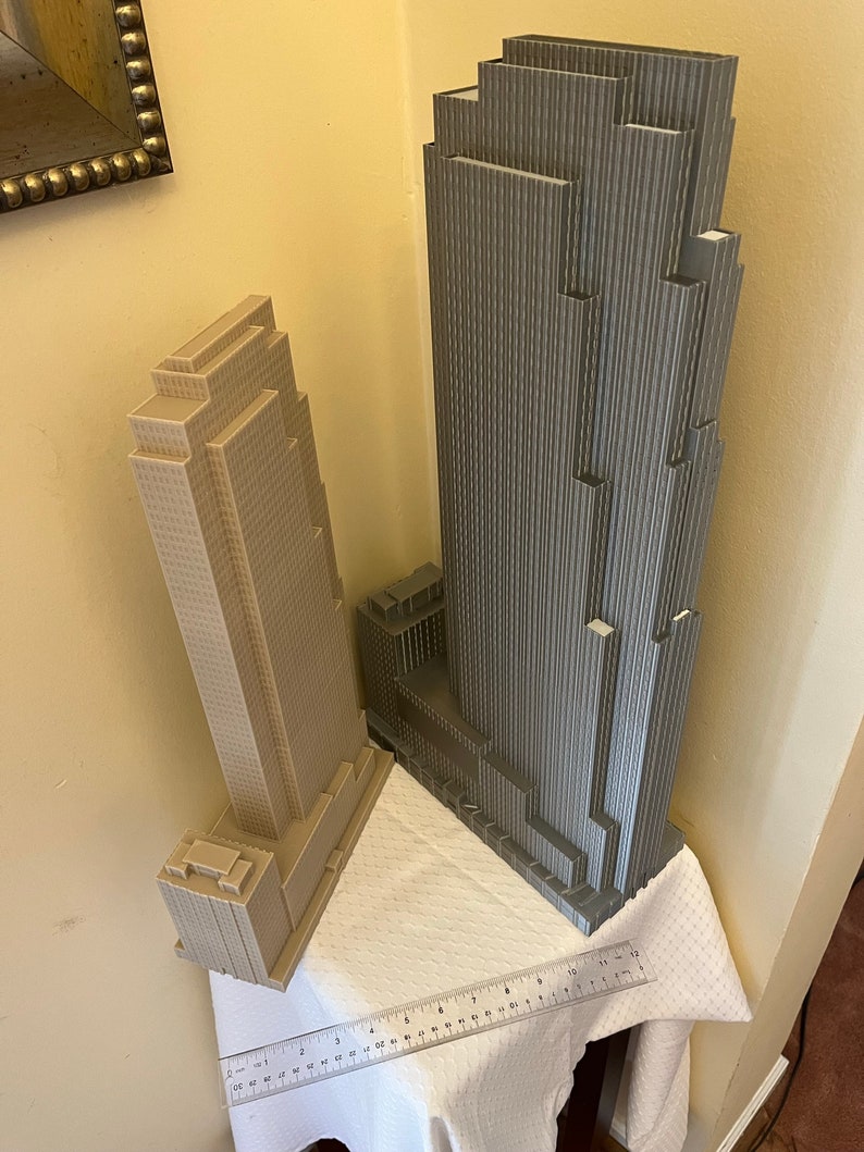 May include: Two 3D printed models of skyscrapers, one is gray and the other is beige. The models are on a white tablecloth with a ruler measuring 12 inches.