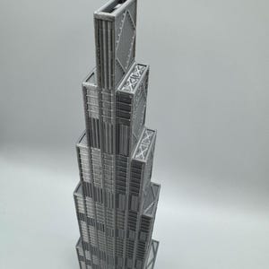 May include: A 3D printed gray model of a skyscraper featuring a stepped, tiered architectural design. The structure displays vertical paneling, recessed window-like textures, and cross-braced geometric patterns on the upper levels of the tower.