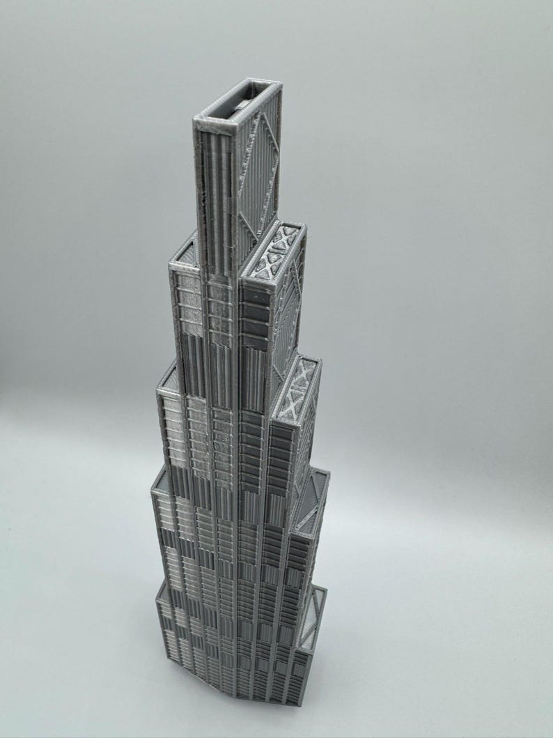 May include: A 3D printed gray model of a skyscraper featuring a stepped, tiered architectural design. The structure displays vertical paneling, recessed window-like textures, and cross-braced geometric patterns on the upper levels of the tower.