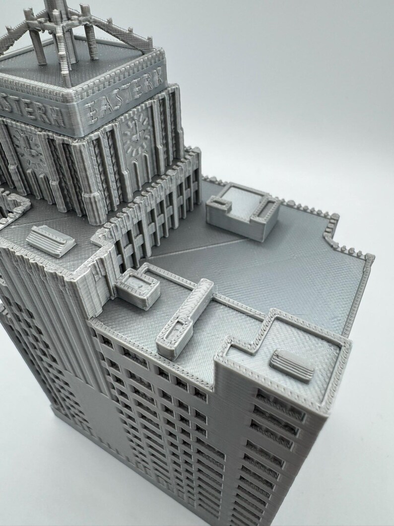 May include: A gray, 3D-printed model of a skyscraper with the word "EASTERN" printed on the side. The building has intricate architectural details, including windows, decorative elements, and a rooftop structure. The model is a detailed representation of a building.