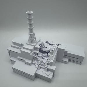 Könnte beinhalten: Ein mattweißes 3D-gedrucktes Modell eines Kernkraftwerks mit einem hohen Entlüftungsschornstein und einem zentralen Bereich, der in einem Zustand strukturellen Einsturzes dargestellt ist, mit verstreuten Trümmerteilen auf dem Dach.